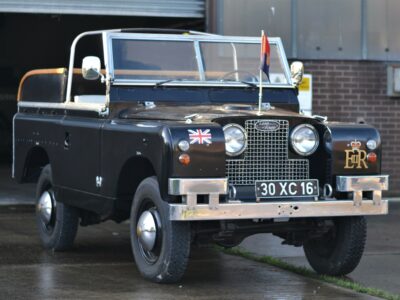Black Land Rover car pictured outside a building with no roof from a diagonal perspective. British Union Jack and Queen Elizabeth II's cypher are printed on the front either side of the number plate and grill, a blue and red flag is on the bonnet