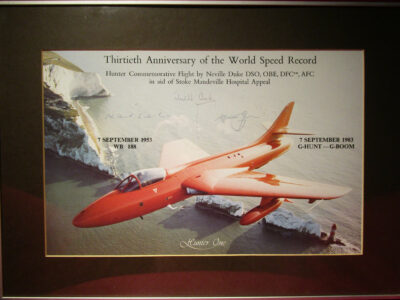 Colour photograph of an orange jet plane flying low over sea cliffs. Photograph is captioned and autographed
